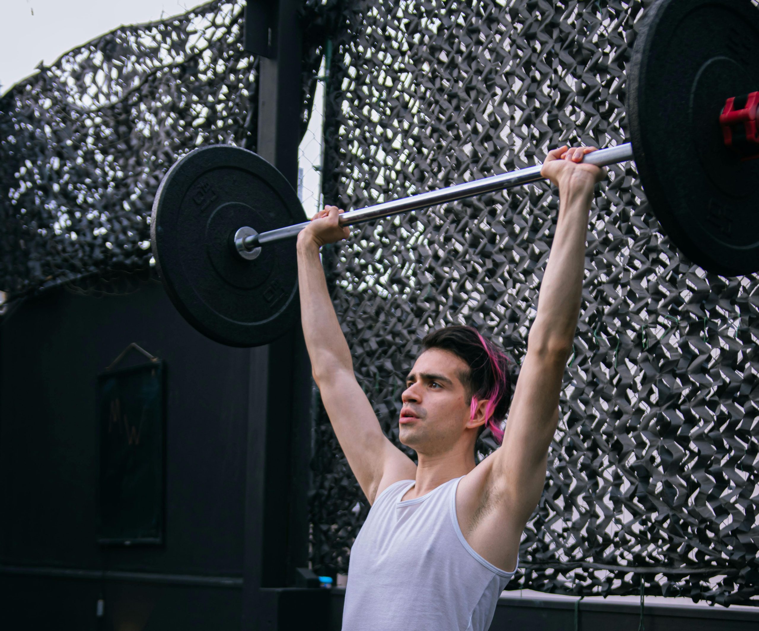 Adult man in white tank top lifting barbell outdoors, showcasing strength and fitness.